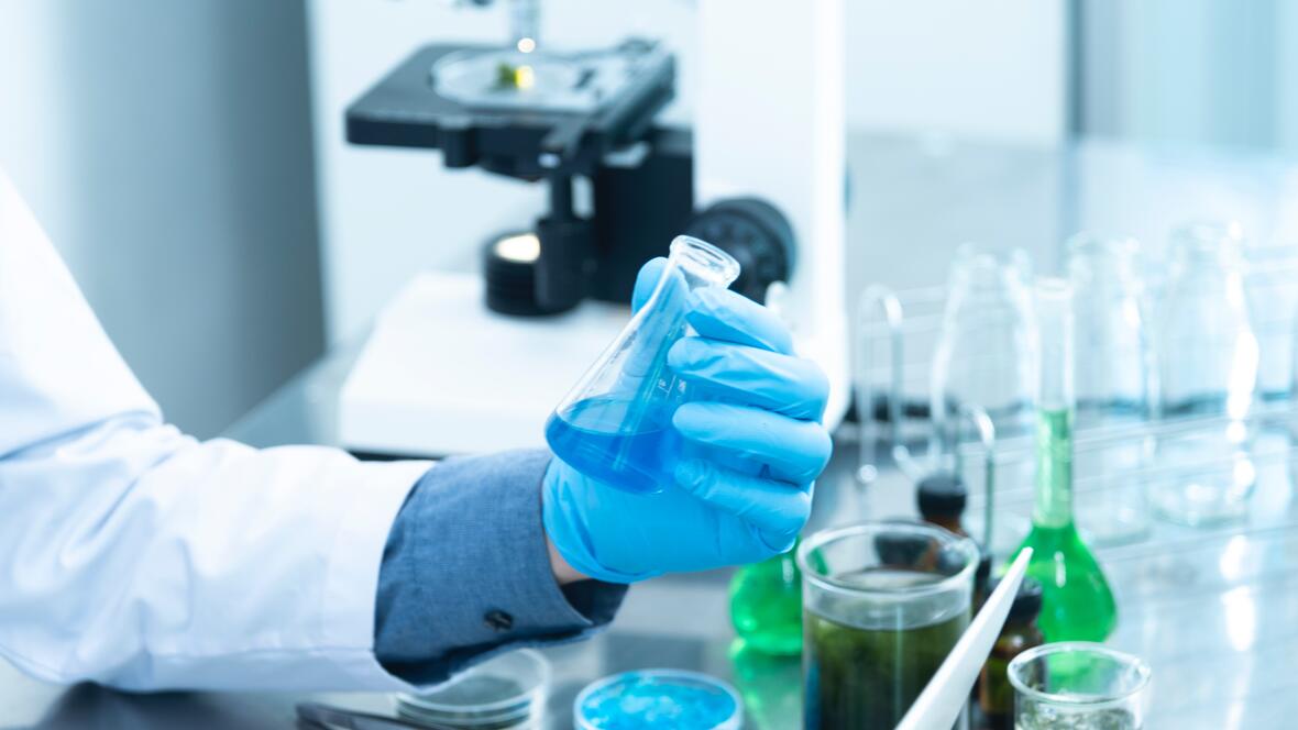 Zu sehen ist der Arm einer Person die einen weißen Laborkittel und hellblaue Labor Handschuhe trägt. In der Hand hält die Person ein Glas, mit blauer Flüssigkeit. Im Hintergrund sind ein Mikroskop und weitere Gefäße zu sehen. Manche sind leer, ein paar enthalten Stoffe in unterschiedlichen Farben.
